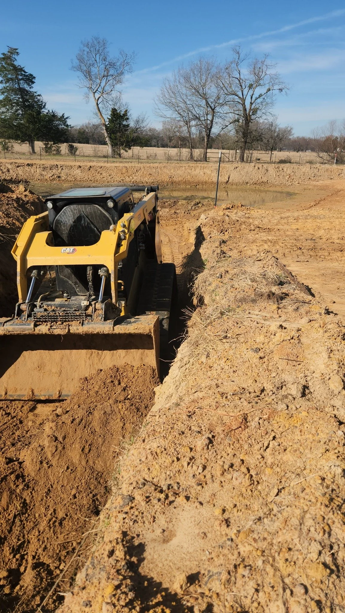 A yellow bulldozer working on a construction site with dug trenches and dirt piles, trees in the background, and a clear blue sky.