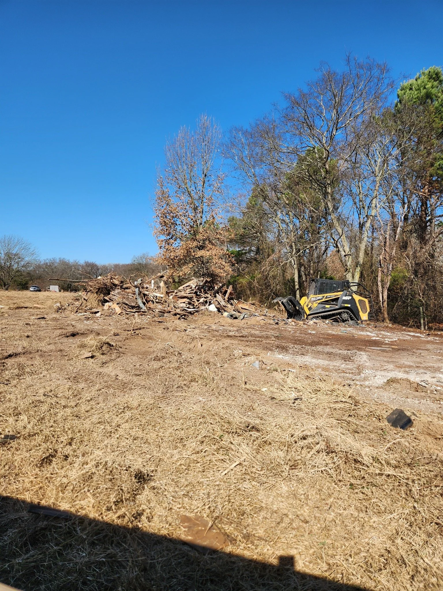 A cleared dirt lot with a small yellow construction vehicle, surrounded by leafless trees under a clear blue sky.
