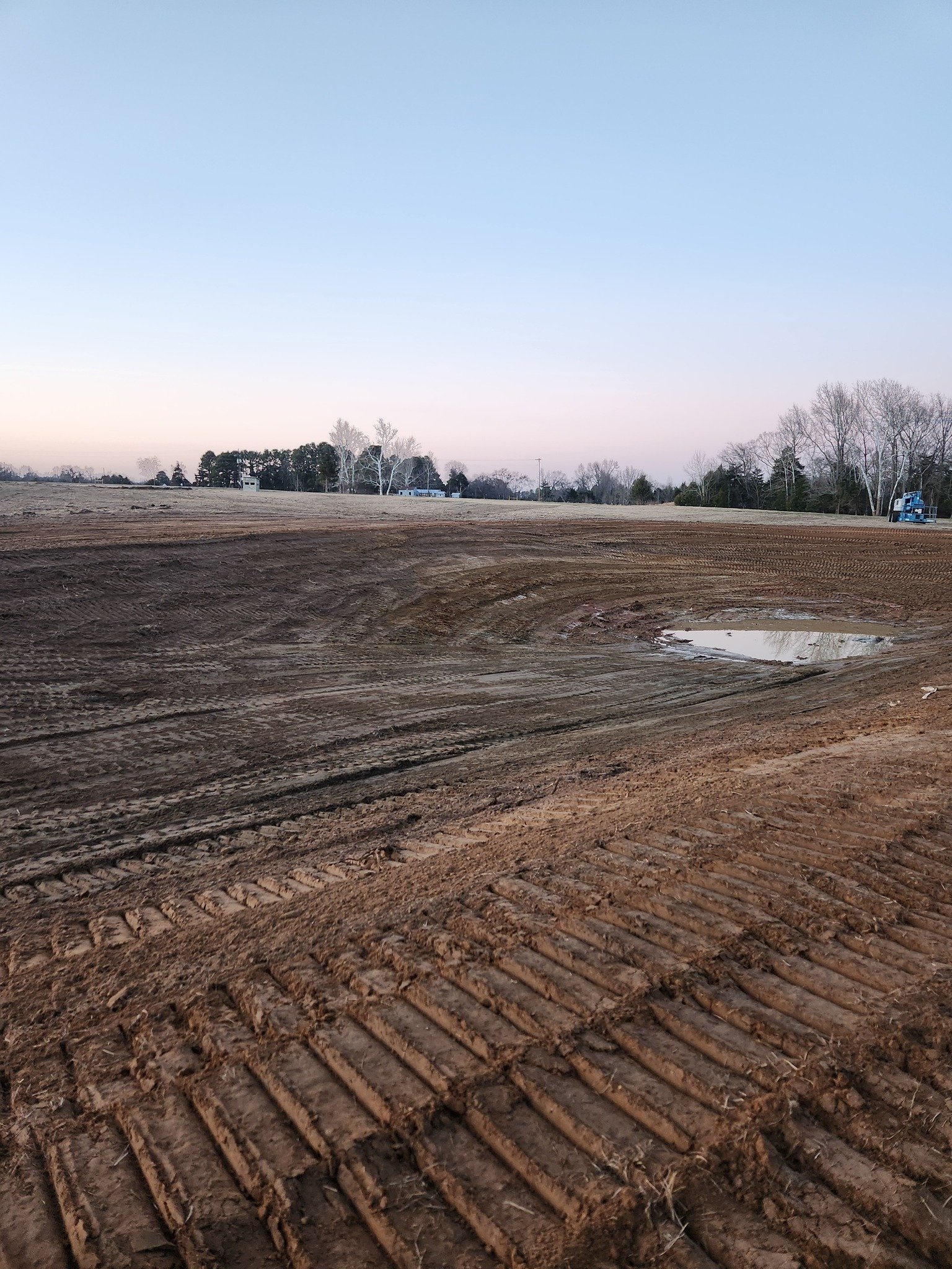 A large open field with freshly tilled soil and visible tire tracks, with a small puddle reflecting the sky, and trees and machinery in the distance under a clear sky at dusk.
