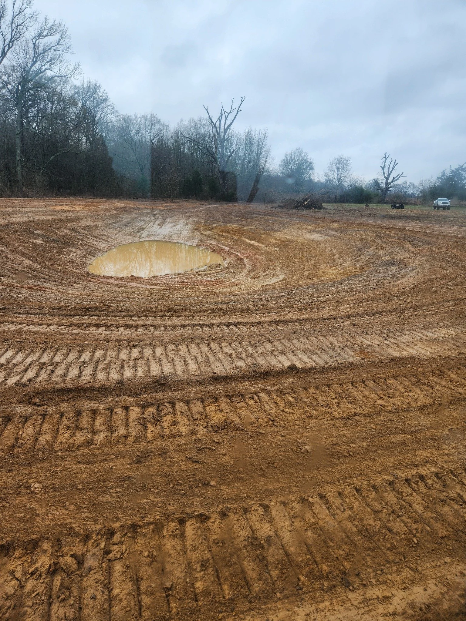 A muddy construction site with tire tracks, a large puddle, leafless trees, and a cloudy sky.
