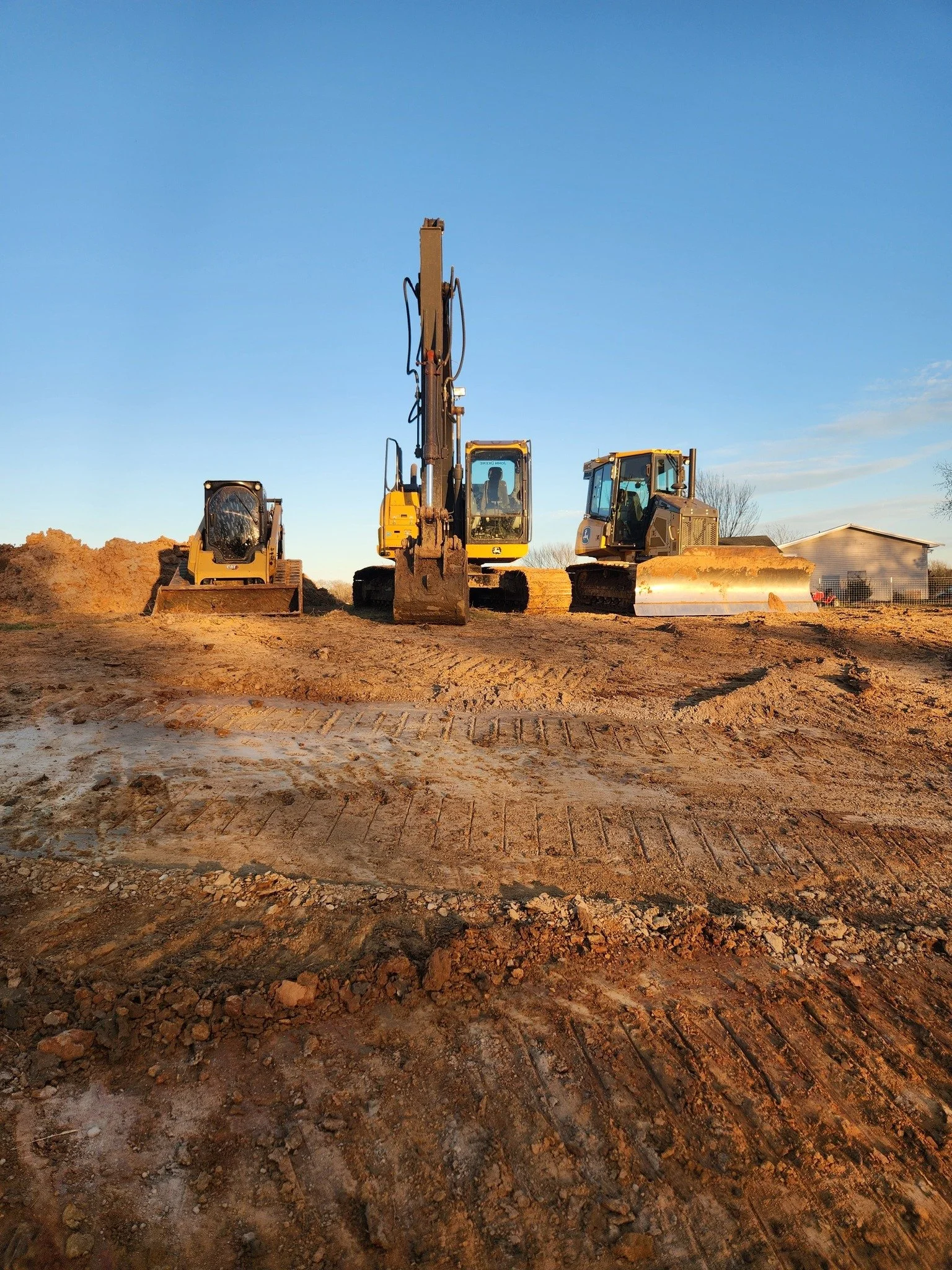 Three construction excavators on dirt ground during sunset with a clear blue sky and a house in the background.