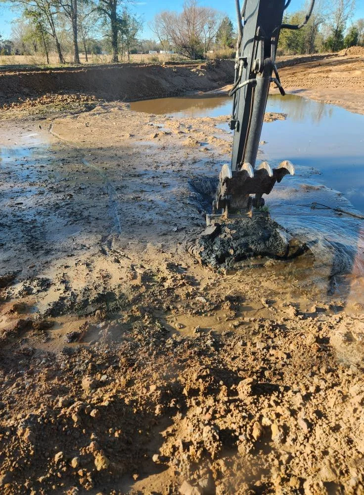 Close-up of an excavator digging in muddy ground near a water puddle with trees in the background.