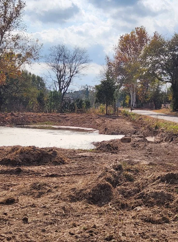 A cleared area with exposed dirt and soil, possibly for construction, with trees and a road in the background under a partly cloudy sky.