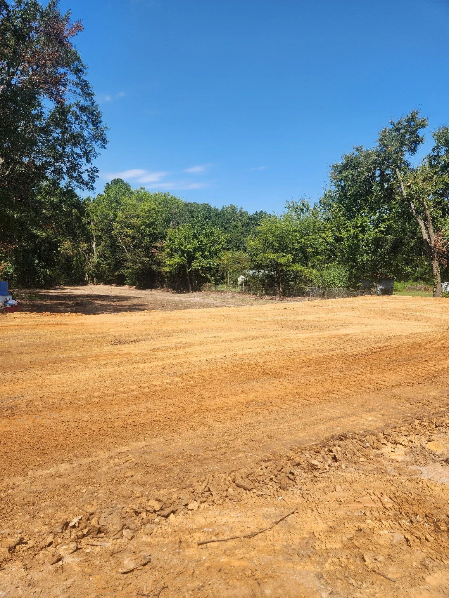 A dirt lot with orange-brown soil, surrounded by green trees under a clear blue sky.