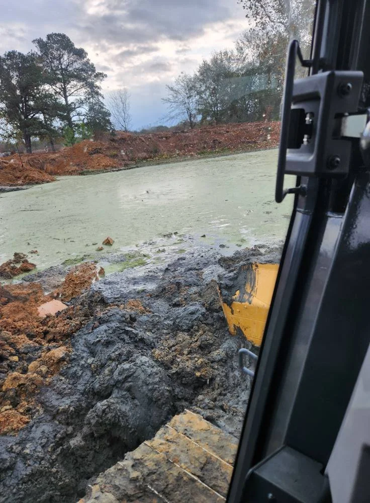 View from an excavator showing muddy earth and a green algae-covered pond with trees and cloudy sky in the background.