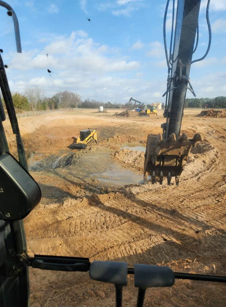 View from inside a construction vehicle showing a large excavator digging at a construction site with dirt and water, under a partly cloudy sky.