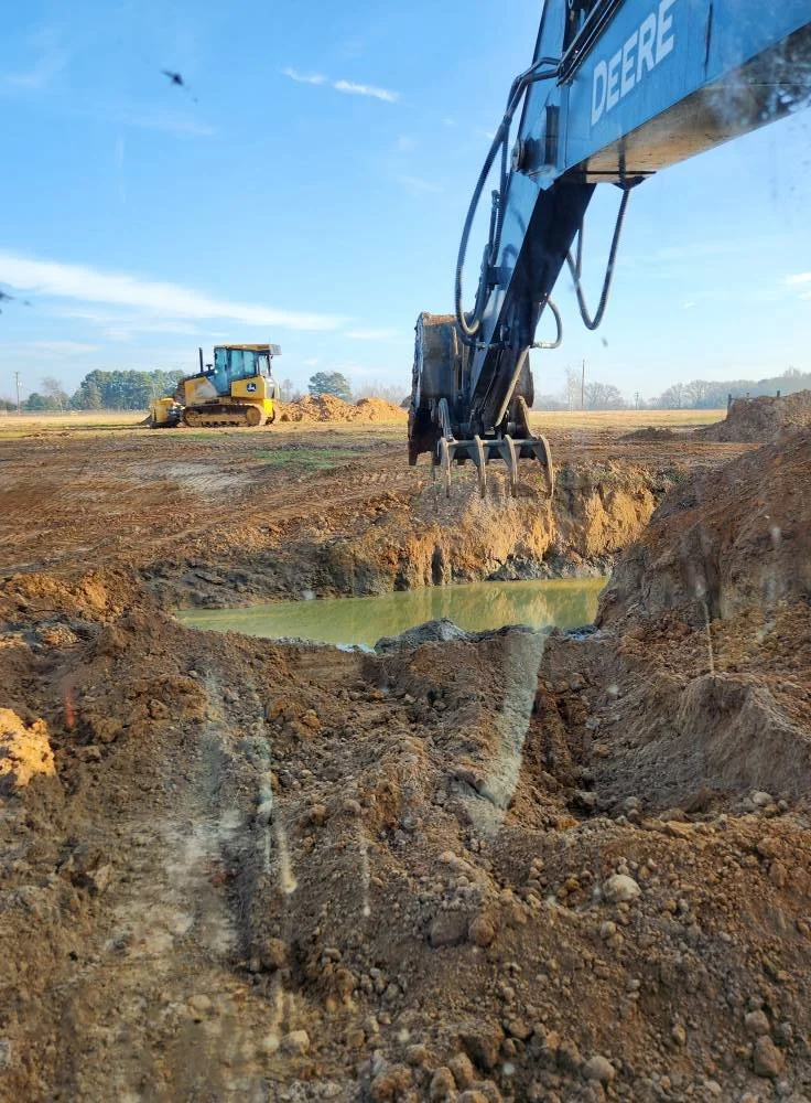 An excavator working on a construction site digging near a pond, with a bulldozer in the background, in a rural area during daytime.