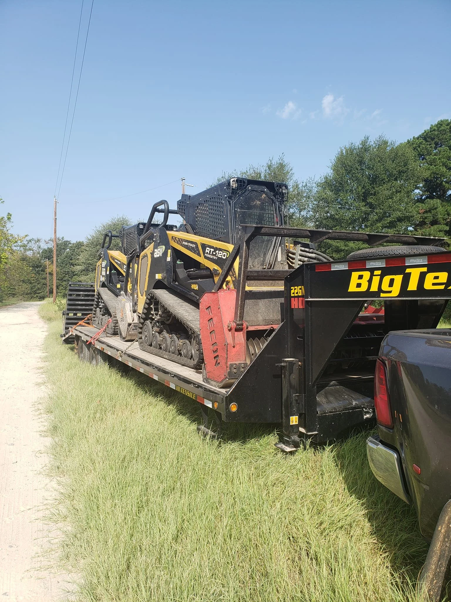 A black flatbed truck carrying two small yellow and black tracked construction machines parked on a grassy roadside with trees and a utility pole in the background.