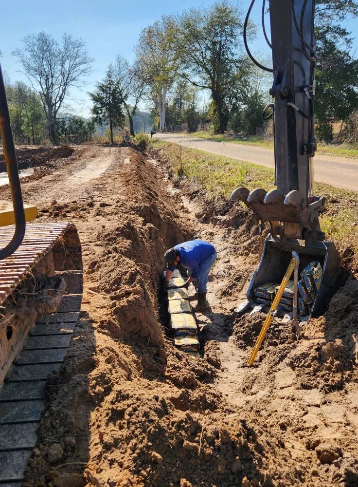 A construction worker in blue overalls working in a trench next to an excavator on a dirt road, with trees and a street in the background.