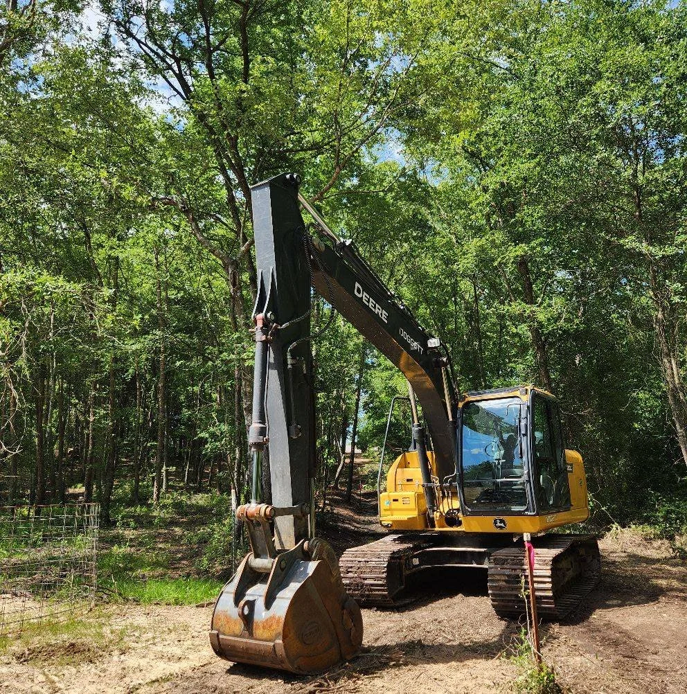 A yellow and black excavator with a large bucket attachment in a wooded area with tall trees and green foliage.