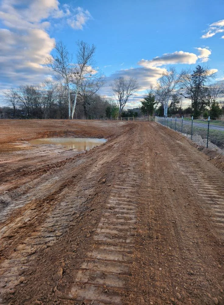 A dirt construction site with tire tracks, a small pond, and a chain-link fence alongside a road with trees and a partly cloudy sky.