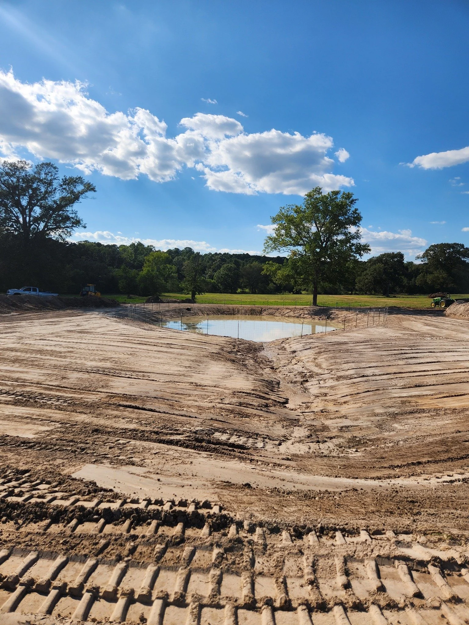 Construction site with a small pond, tire tracks in the dirt, a tree in the background, and construction equipment on the side, under a blue sky with clouds.