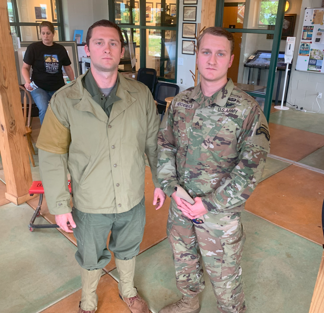 Two men in military uniforms stand side by side indoors, with a woman in casual clothing in the background.