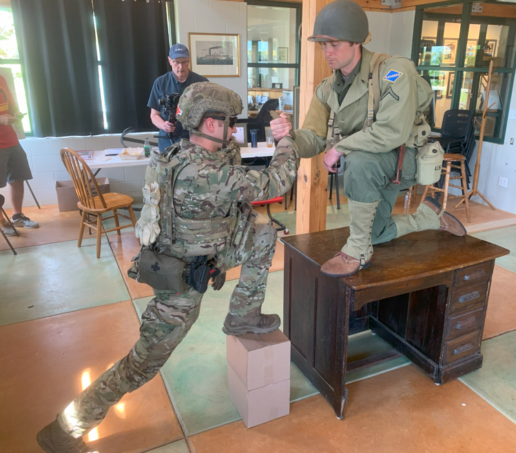 A soldier in camouflage gear and helmet arm wrestling a man in a military uniform with a green helmet, standing on a desk. Two other men are observing, one in a blue shirt and hat, and another in an orange shirt, inside a room with wooden furniture a