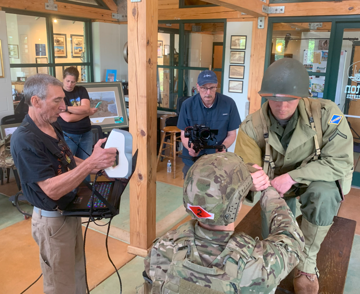 A military soldier in uniform and helmet is sitting at a table, receiving a vaccination shot from a person wearing a tan military uniform and helmet. Two men in civilian clothing are observing, one holding a camera and the other taking notes, in a ro