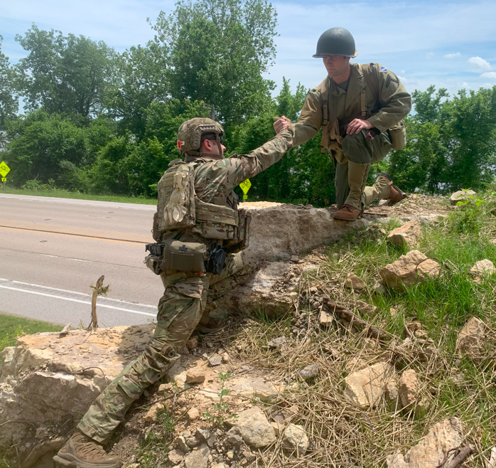 A soldier in camouflage uniform shaking hands with a police officer who is kneeling on a rock by the roadside, with green trees and a cloudy sky in the background.
