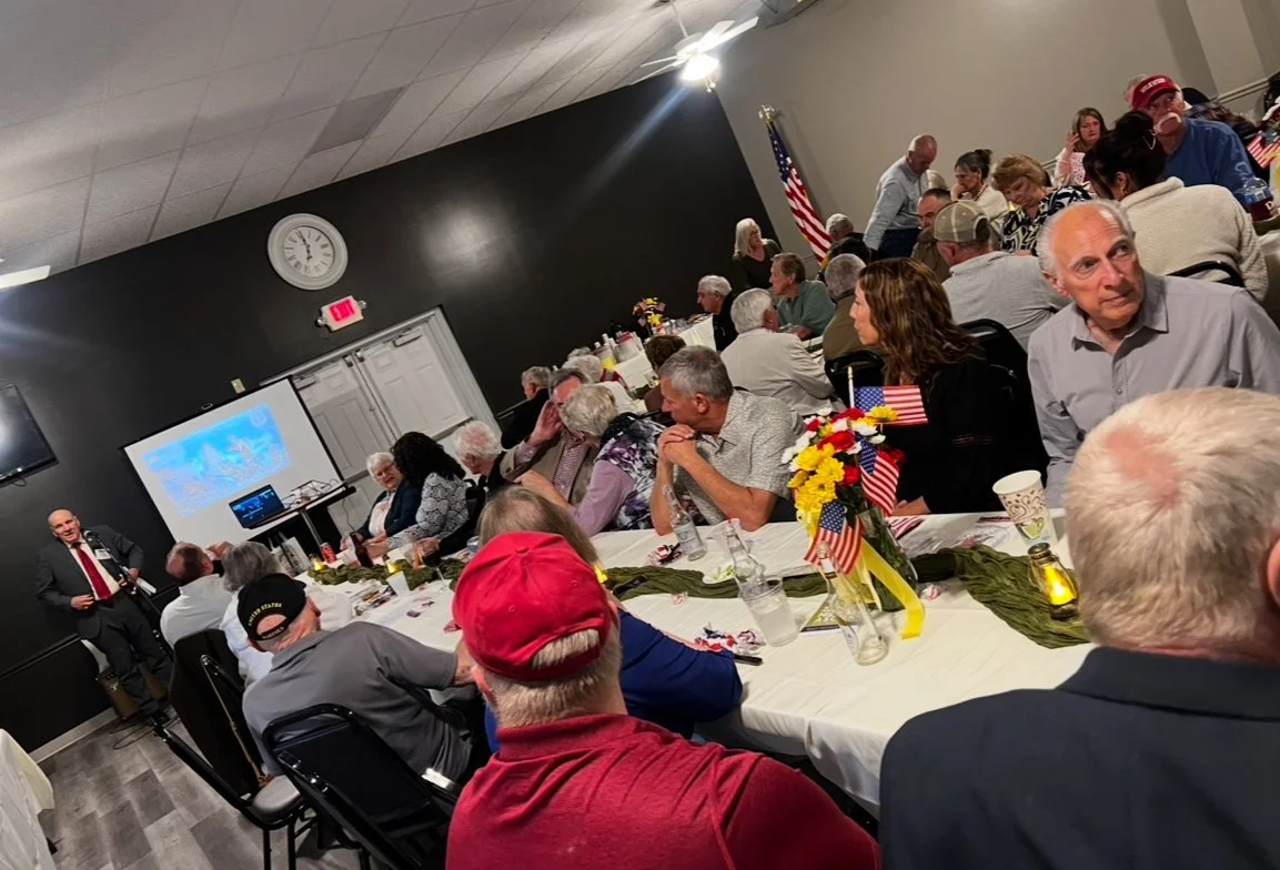A large indoor gathering with multiple people seated at decorated tables, some with American flags, during a patriotic event.