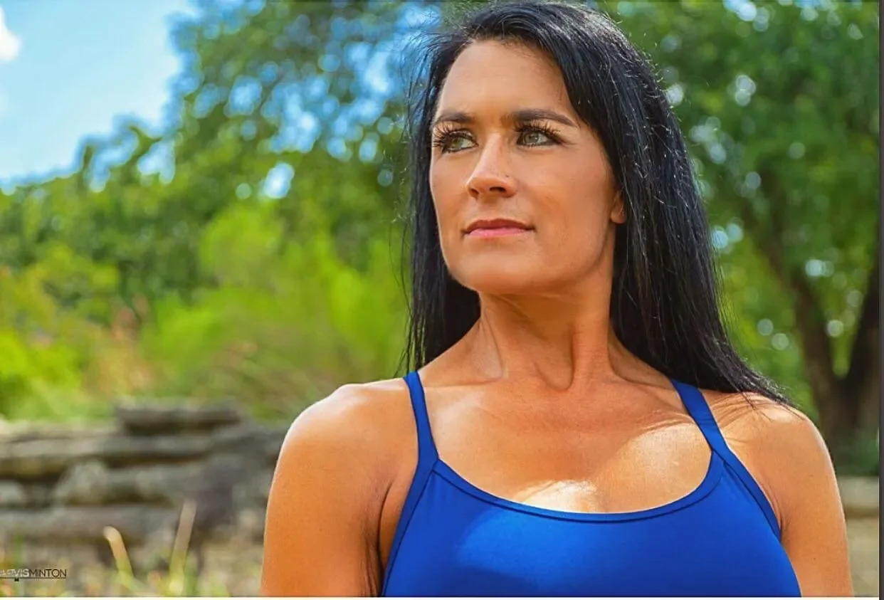 A woman with long black hair, wearing a blue tank top, standing outdoors with green trees in the background.