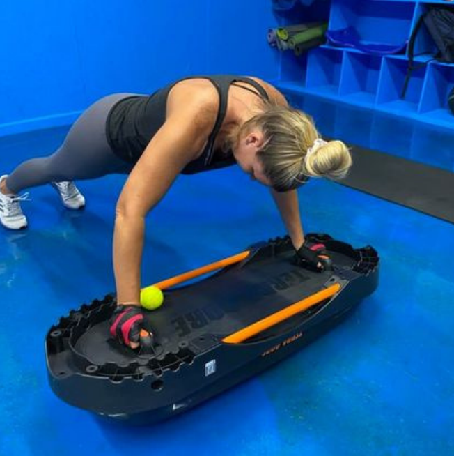A woman exercising on a fitness slide board in a gym class, with a small yellow ball placed under her right hand.