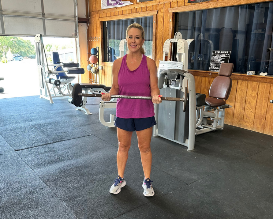 Woman in workout clothing holding a barbell in a gym with exercise equipment and wooden walls.