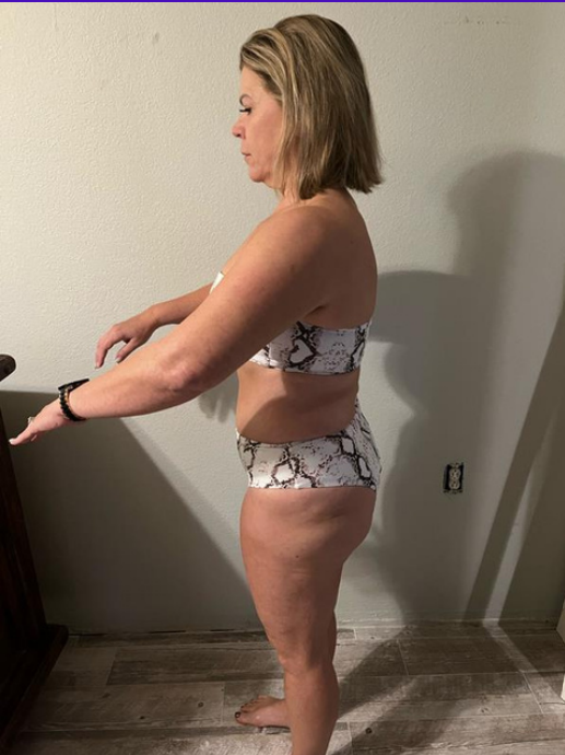 Side view of a woman standing in minimal clothing with her arms stretched forward, in a room with a plain wall and wooden floor.