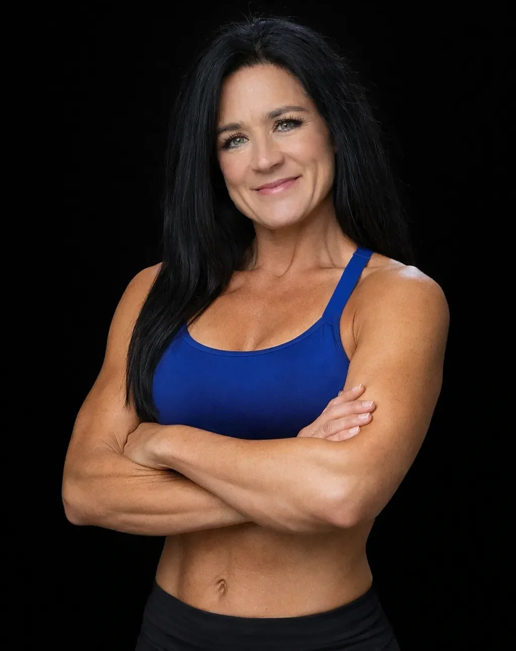 A fit woman with long black hair wearing a blue sports bra and black pants, standing with her arms crossed and smiling against a black background.