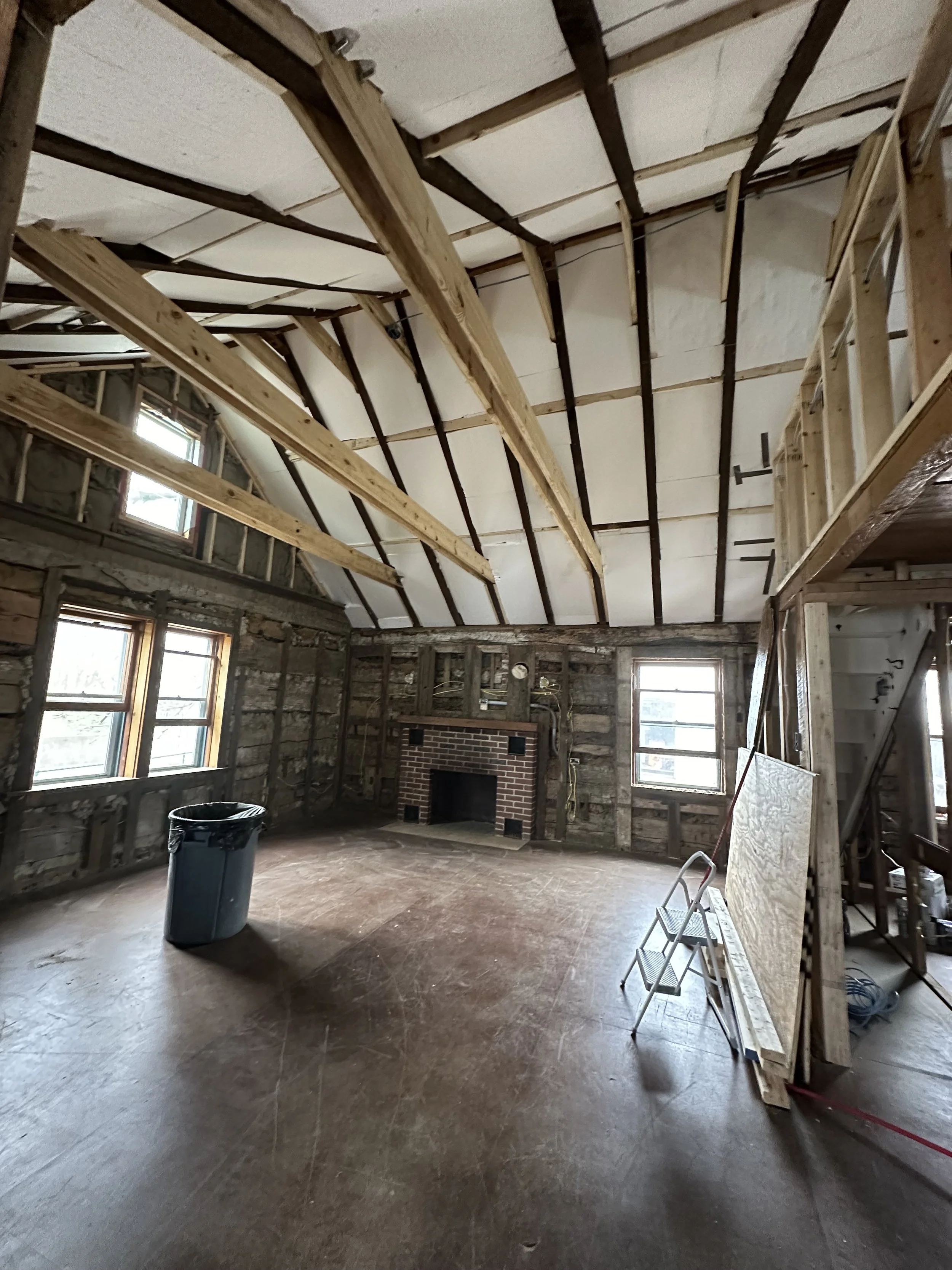 Interior of a room under renovation with exposed wooden beams, brick fireplace, windows, and construction materials.