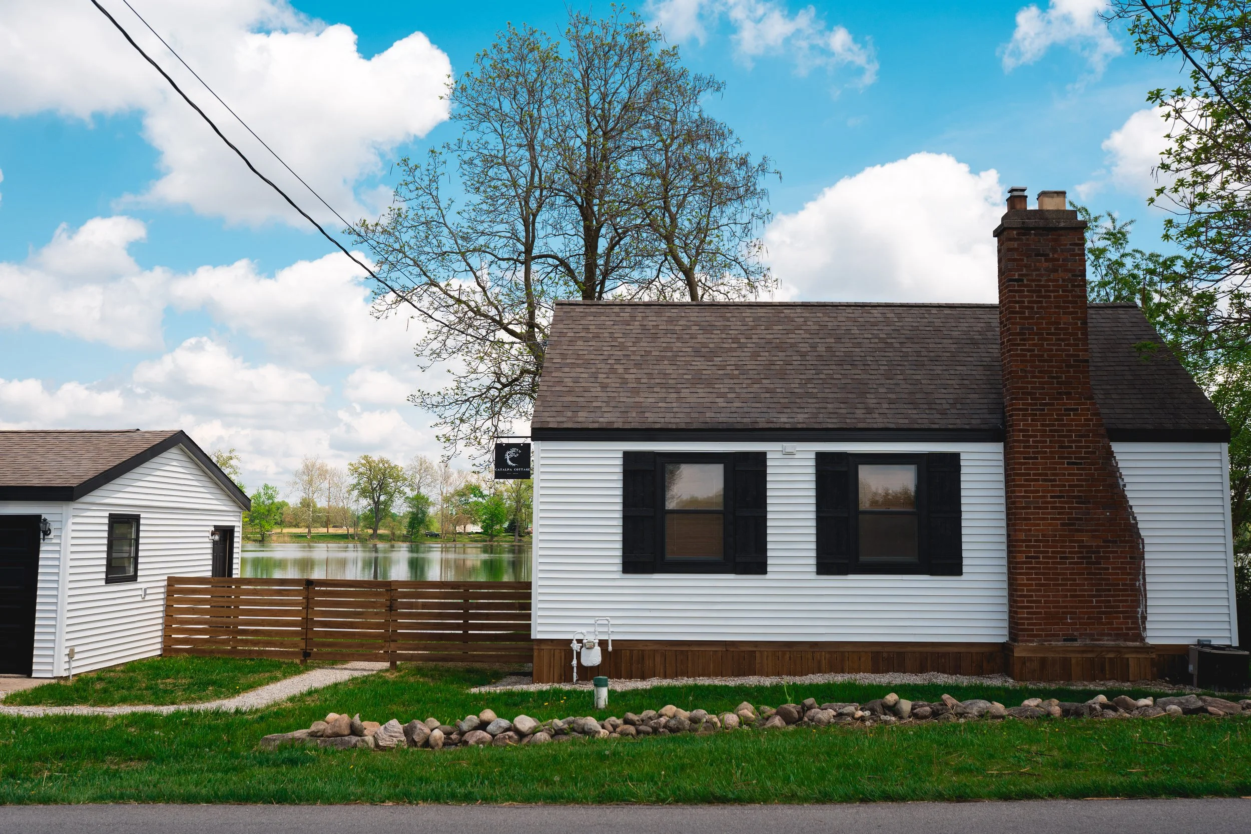A white house with black window shutters, a brick chimney, and a brown roof, next to a smaller white building, with a wooden fence and a lake in the background, under a blue sky with scattered clouds.
