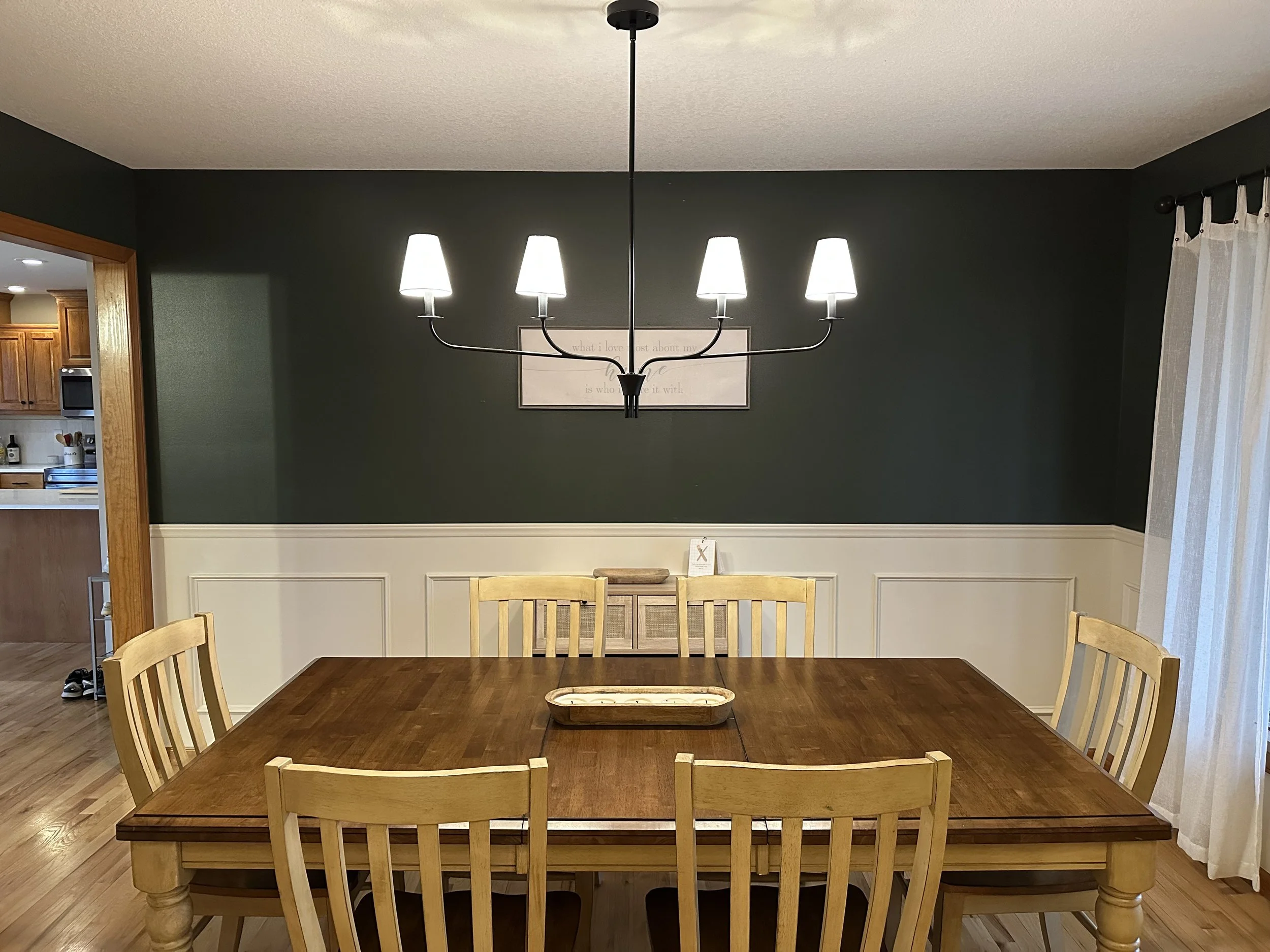 A dining room with a wooden table and six matching chairs. There is a chandelier with five white lampshades hanging above the table. A wall with dark green paint and white wainscoting is in the background. There is a framed quote on the wall and a curtain on a window to the right.