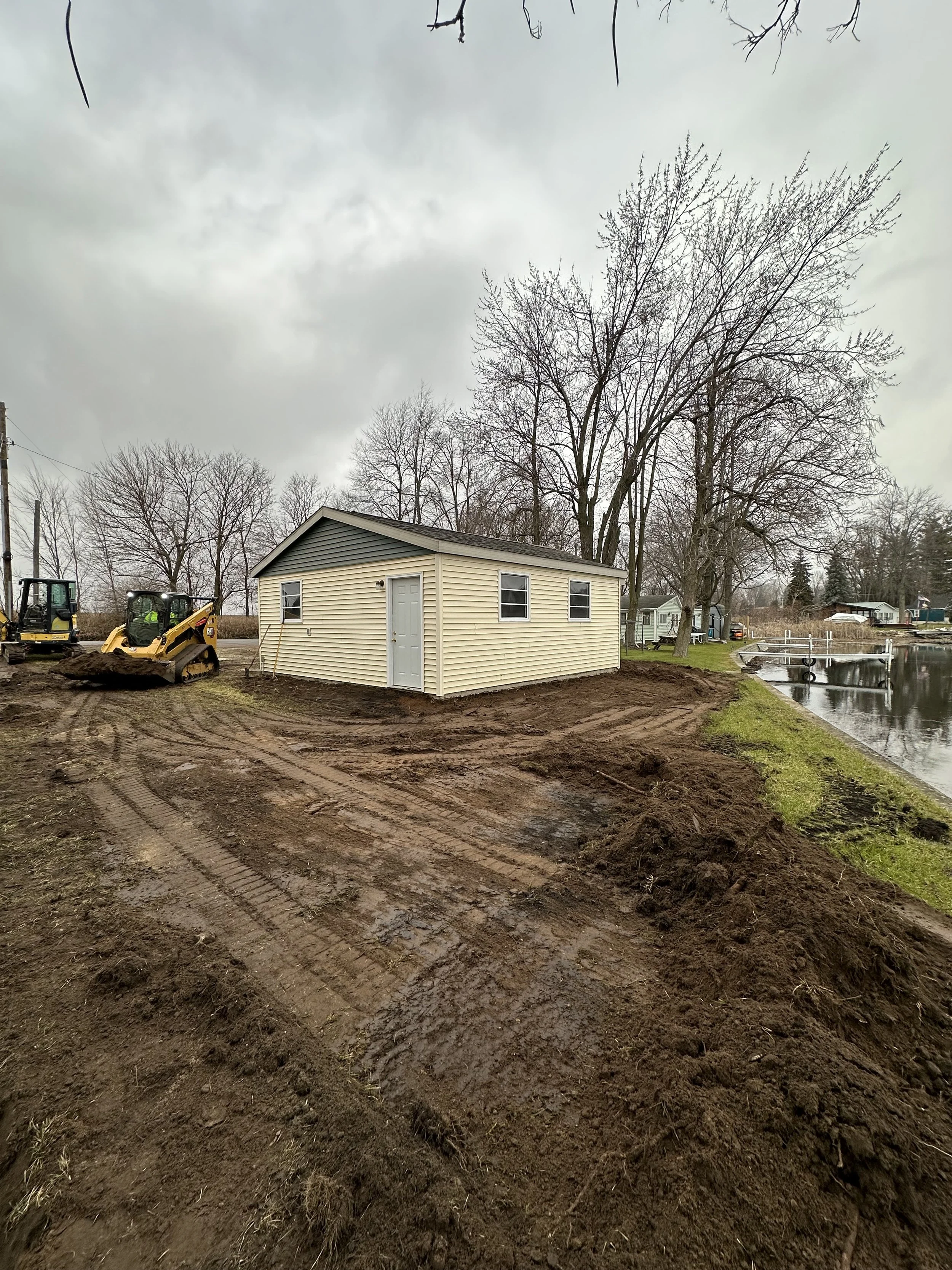 A small beige building with double windows and a door is situated on muddy ground near a body of water, with construction equipment and bare trees in the background under a cloudy sky.