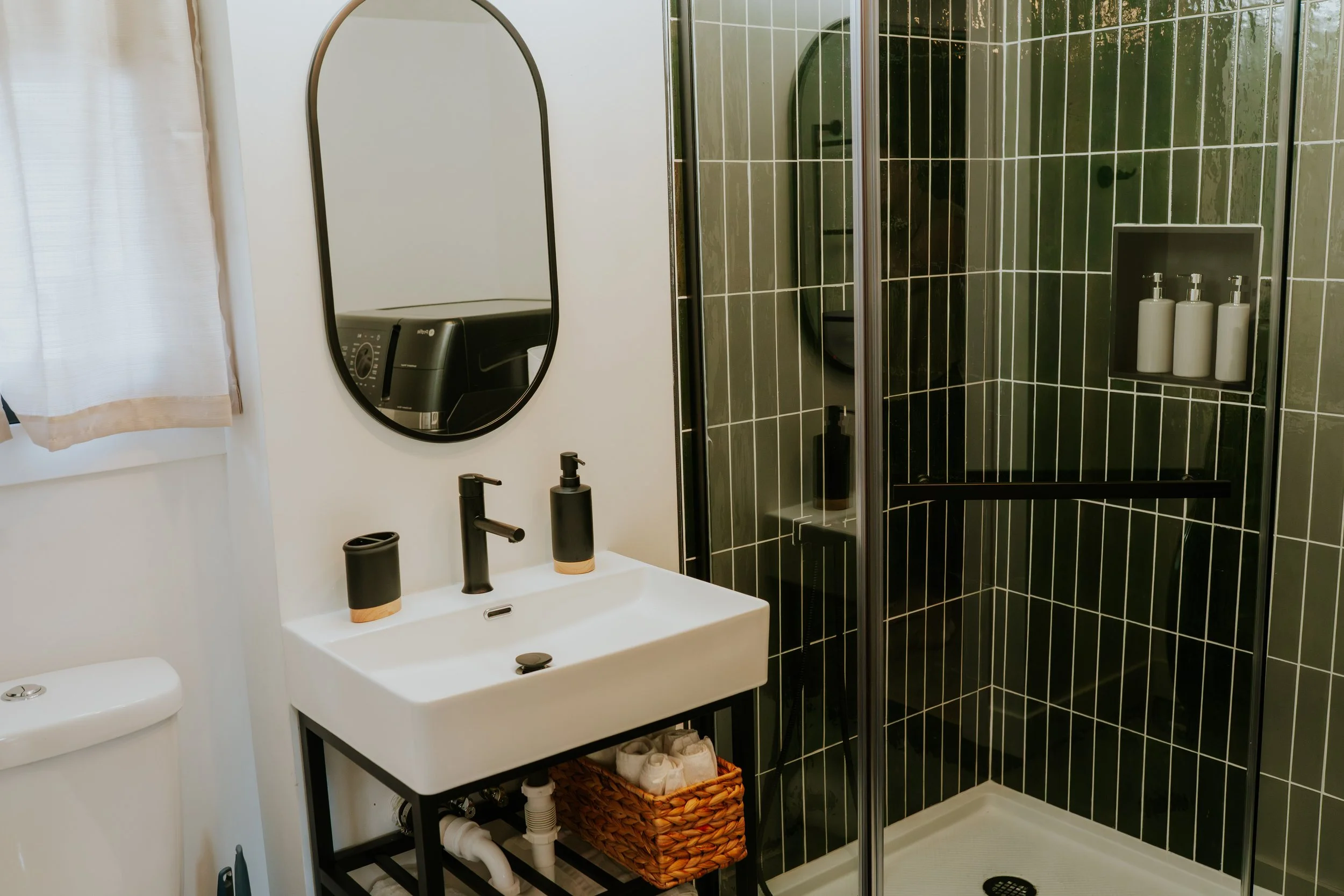 Modern bathroom with black fixtures, a white square sink, a mirror, a toilet, and a black-tiled shower with transparent door and built-in shelves holding toiletries.