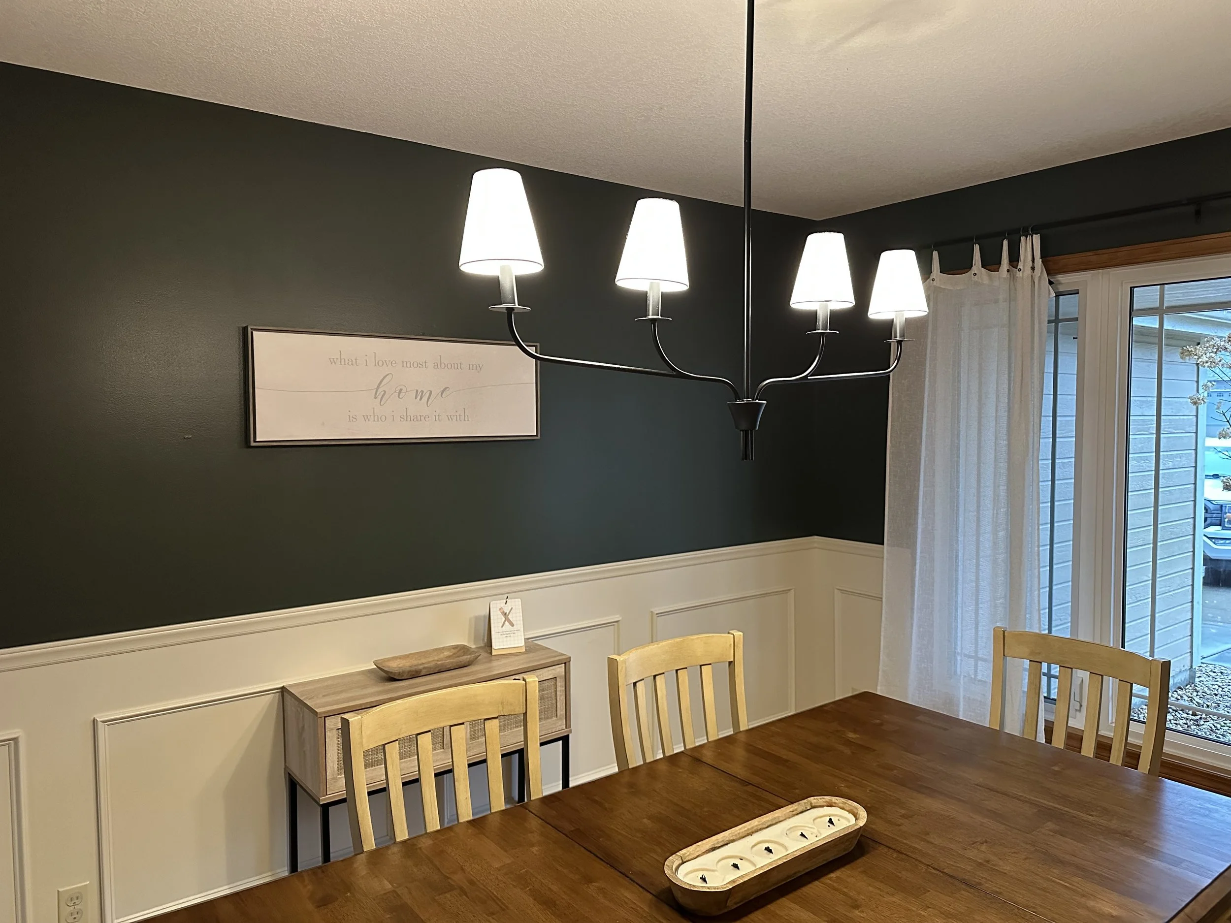 A dining room with a wooden table, wooden chairs, a hanging chandelier with five white lampshades, a small wooden sideboard, and a window with curtains showing an outside view.