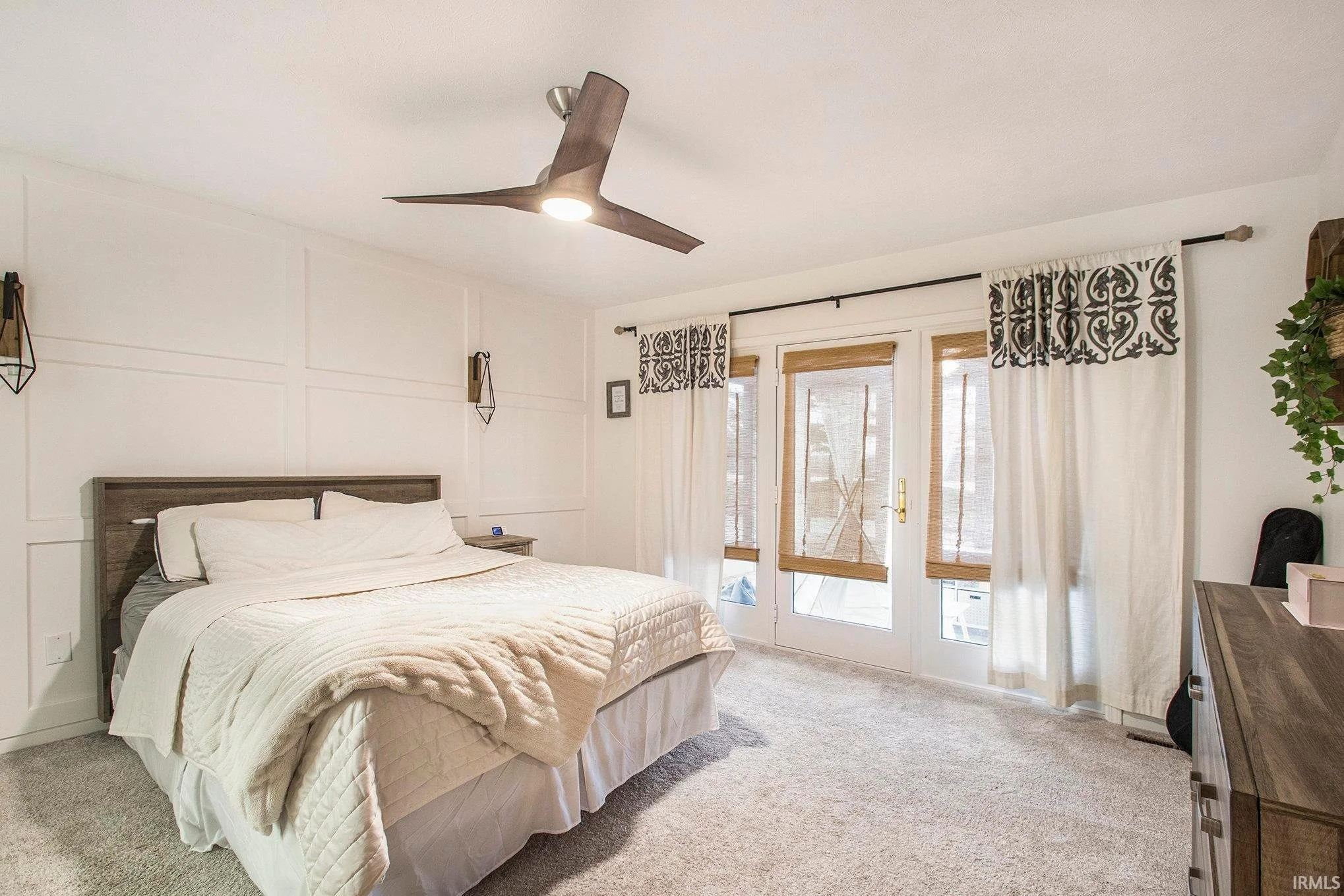 Comfortable bedroom with a large bed, beige bedding, and a ceiling fan with wooden blades, illuminated by a light. The room has windows with bamboo shades and black-and-white patterned curtains, and has a light-colored carpet. There are wall-mounted lighting fixtures and a wooden dresser.