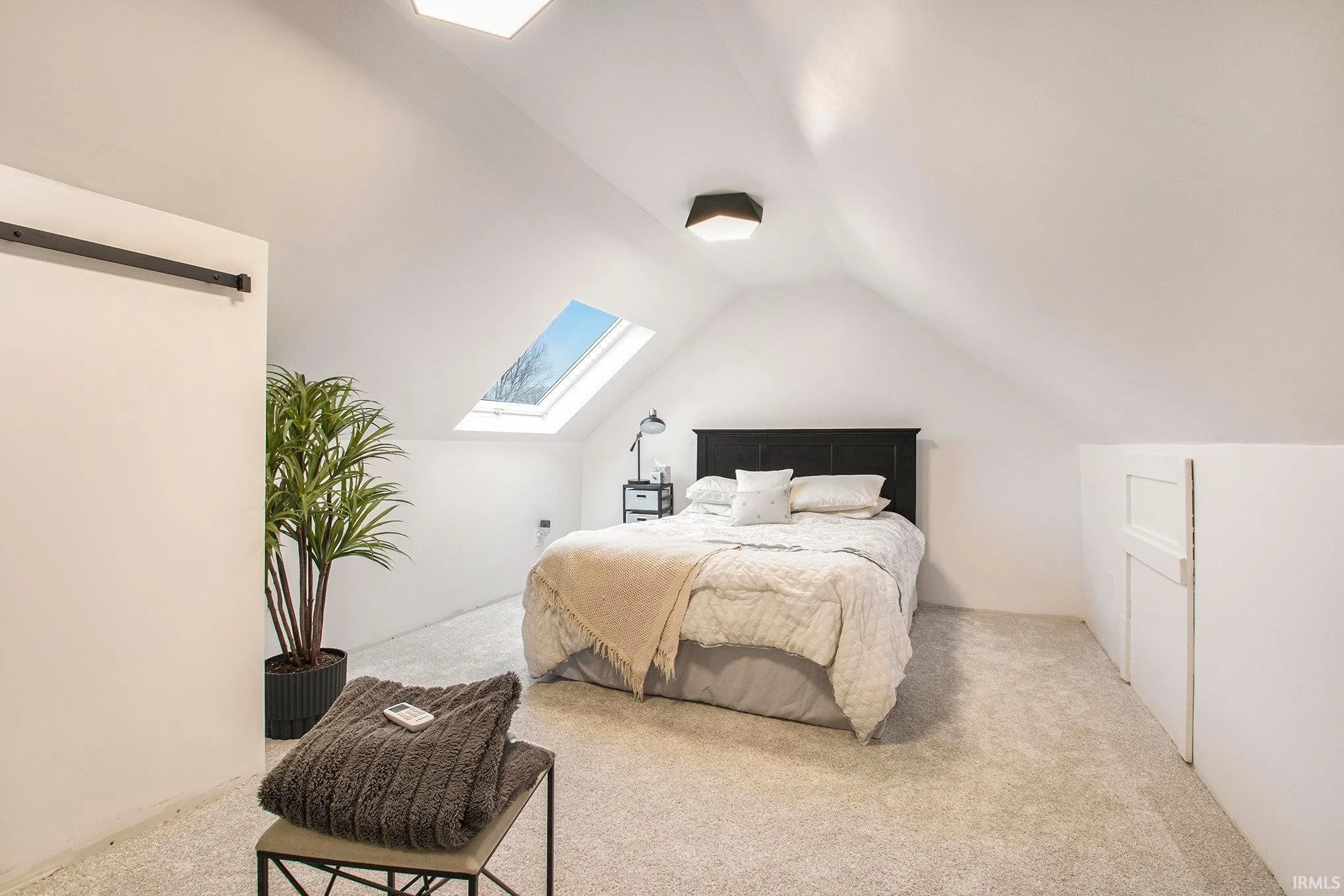 A cozy bedroom with a sloped ceiling, a skylight window, a black headboard, white bedding, a small black nightstand with a lamp, a potted plant, and a gray blanket on a chair, all inside an attic space.