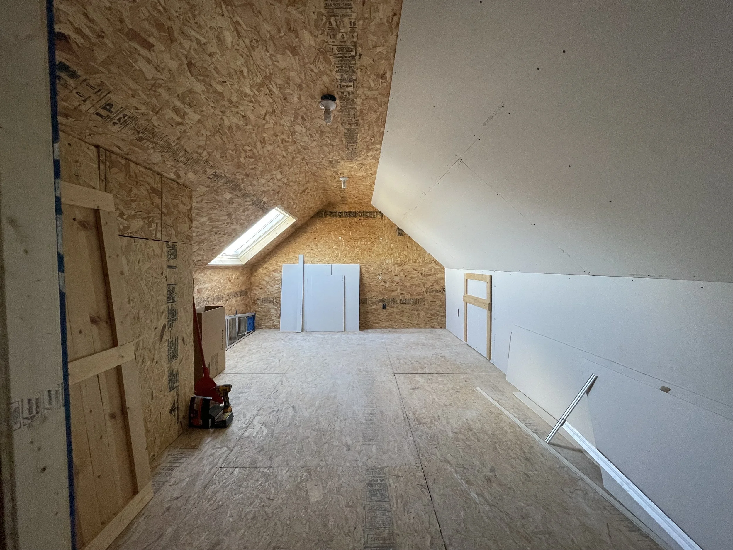 An attic space under construction with plywood and drywall walls, a skylight window, and construction tools and materials on the floor.