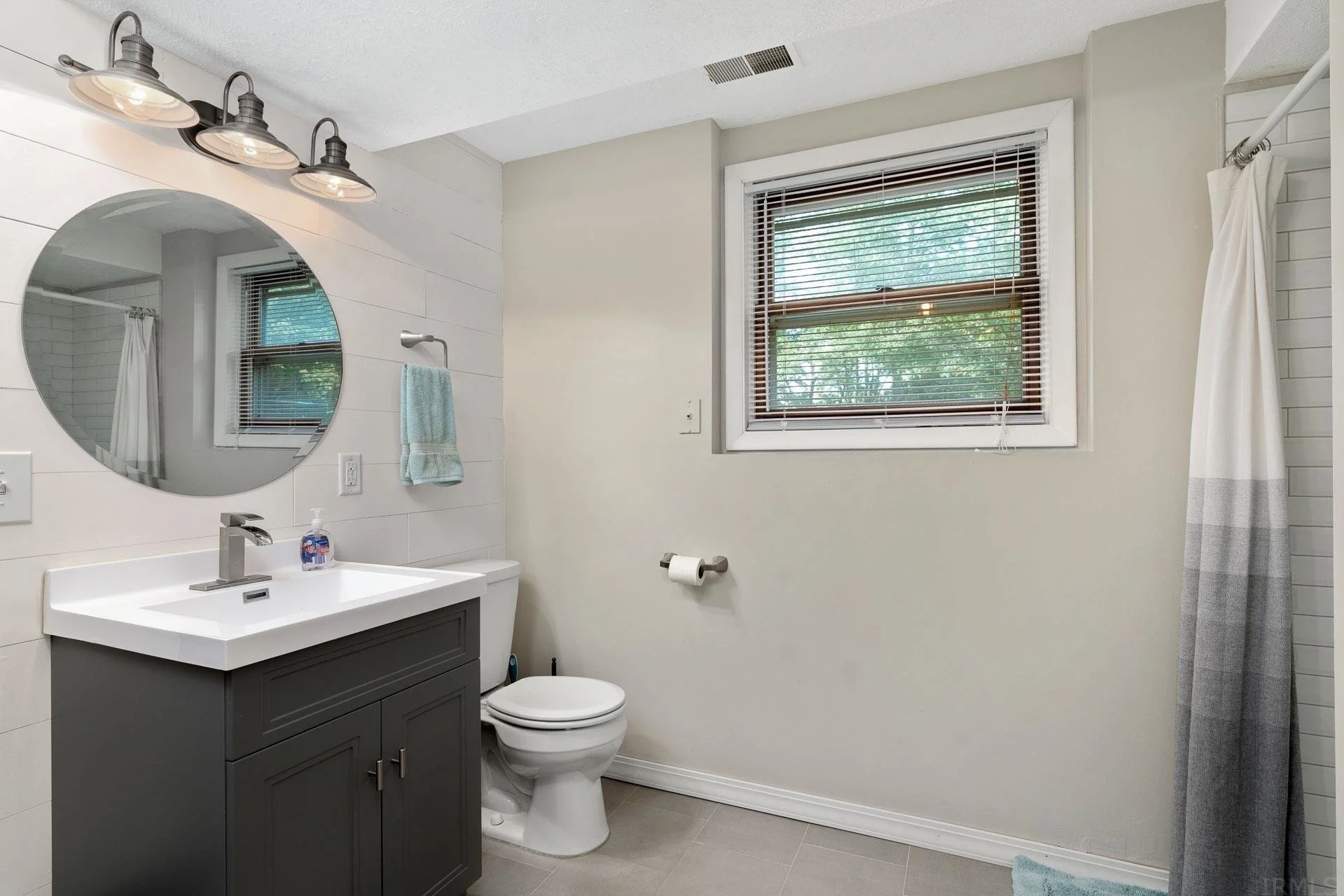 A bathroom with a modern vanity, round mirror, wall-mounted light fixtures, toilet, window with blinds, and a shower curtain.