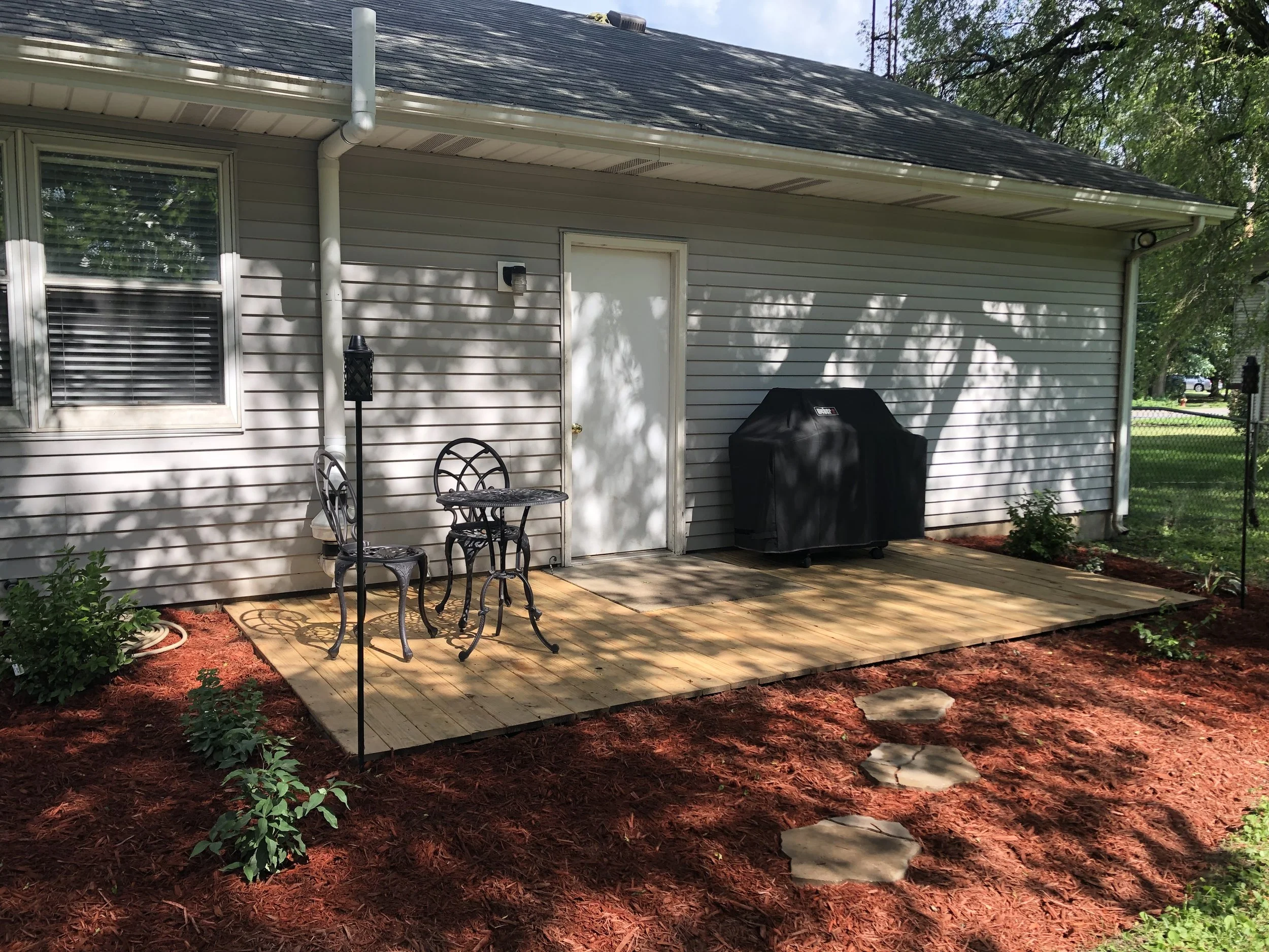 Backyard patio with a wooden deck, black outdoor furniture, barbecue grill, and flower beds with red mulch.