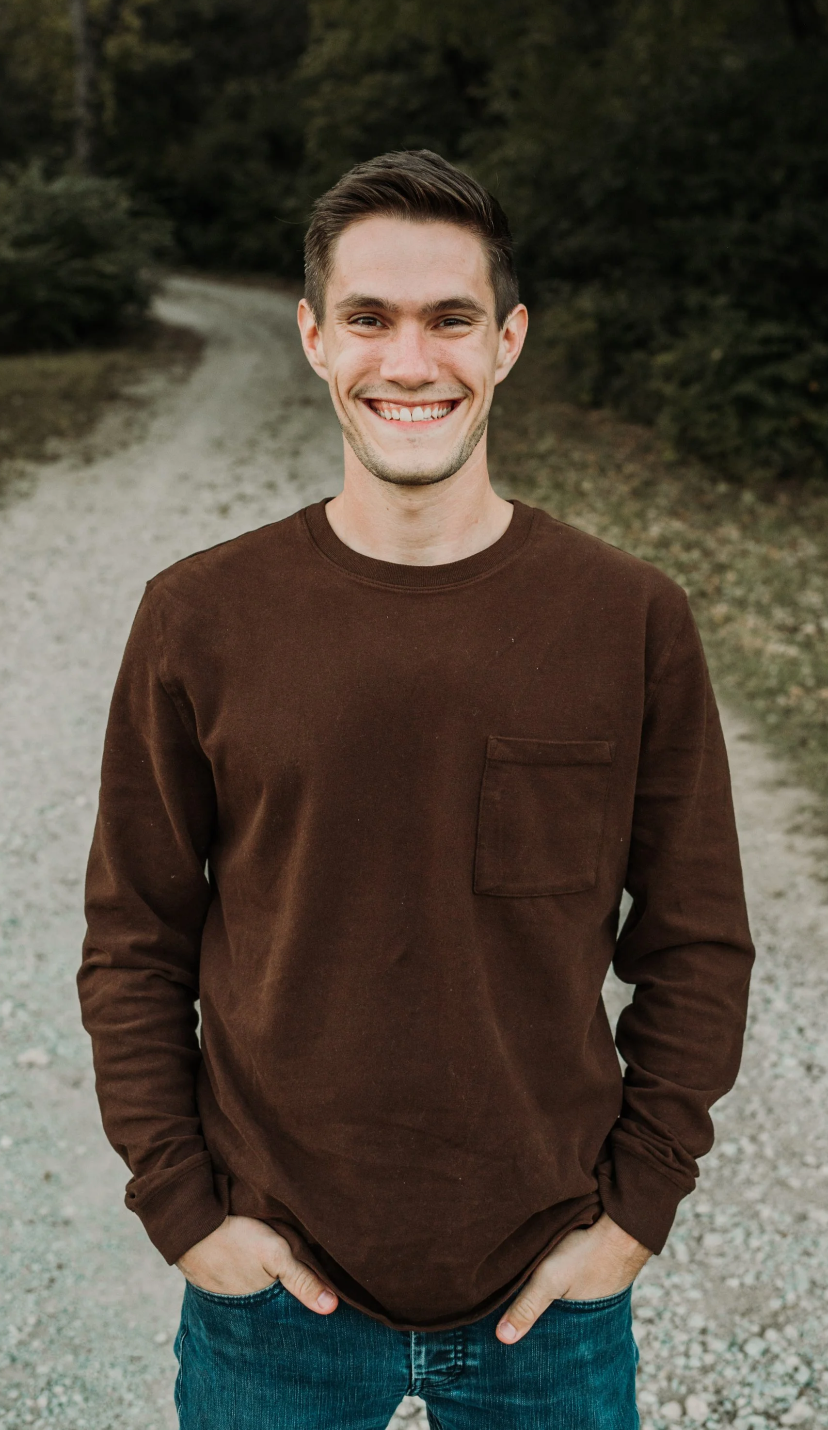 A young man smiling outdoors on a gravel path with trees in the background, wearing a brown long-sleeve shirt and blue jeans.