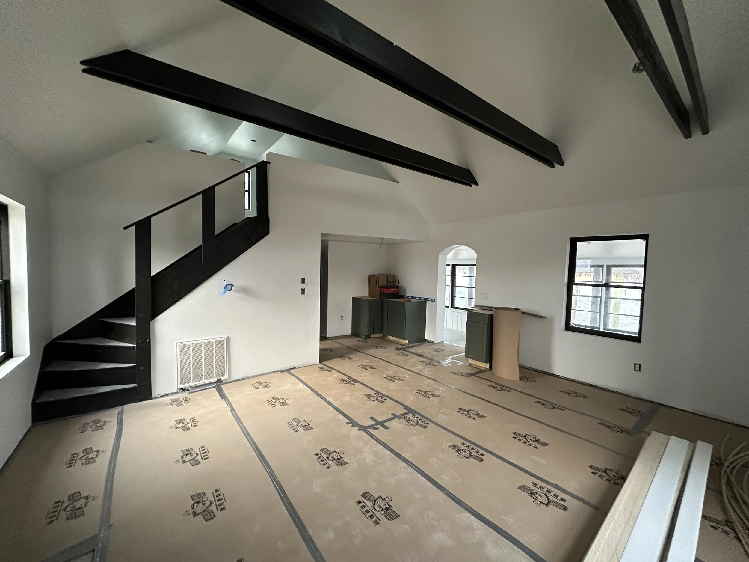 Interior of a house under construction with white walls, black ceiling beams, a black staircase, windows letting in natural light, and flooring covered with protective cardboard.