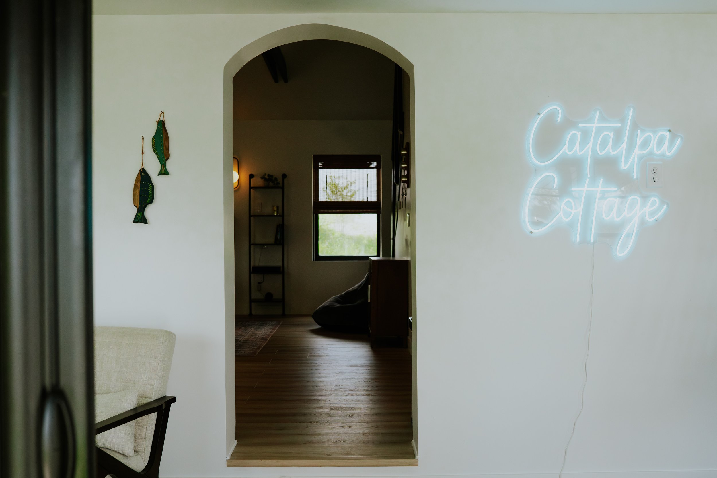 Interior view of a room with a white wall, a neon sign reading 'Catalpa Cottage' on the right, and decorative fish wall hangings on the left. Through a doorway, a room is visible with wooden flooring, a window, and a shelf with decor items.