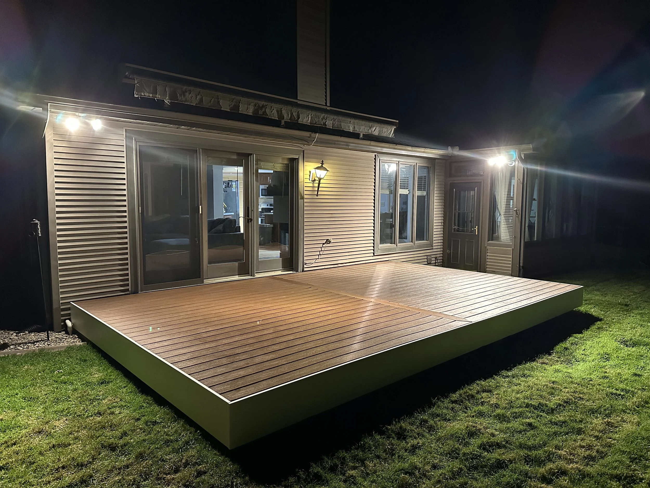Backyard deck illuminated at night, attached to a house with sliding glass doors and windows, outdoor lights, and a grassy lawn.