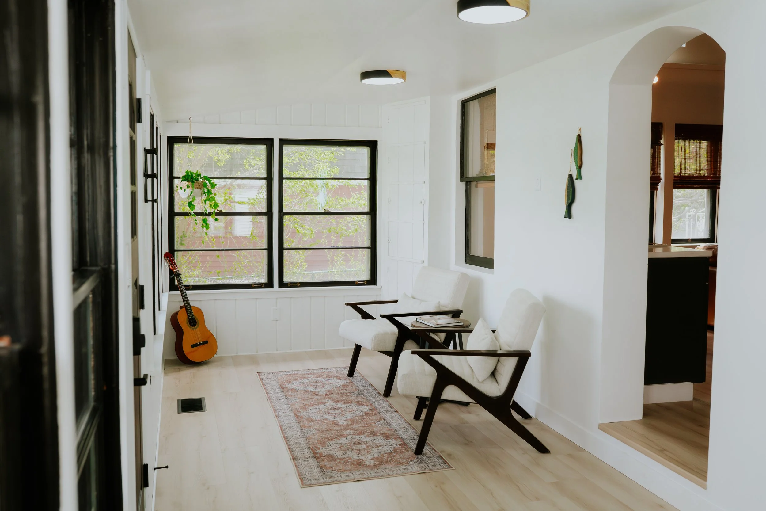 Bright living room with white walls, wooden floor, large black-framed windows, two white armchairs with dark wooden frames, a small rug, an acoustic guitar leaning against the wall, and a hanging potted plant.
