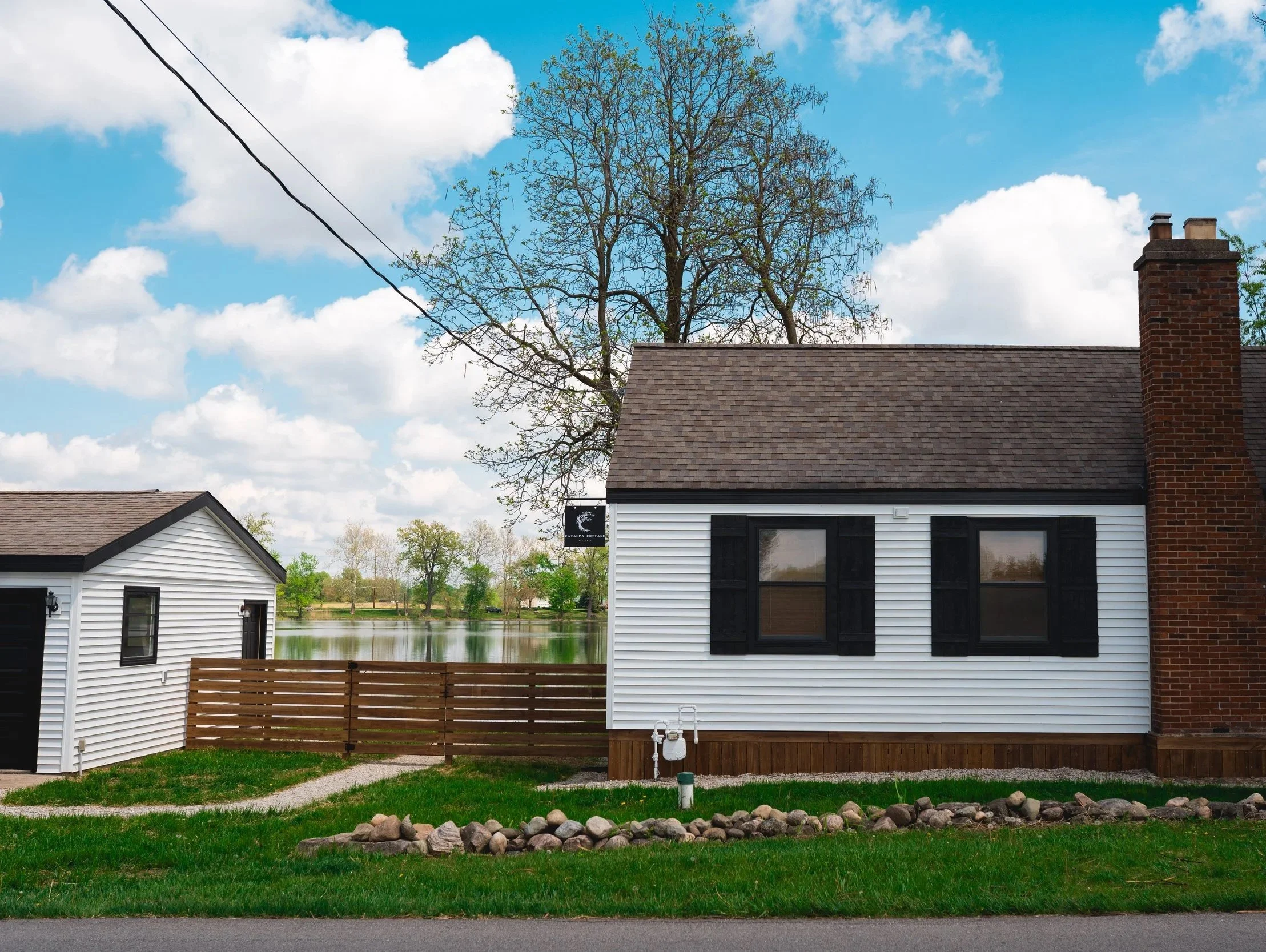 A white house with black shutters and a brick chimney, next to a smaller white shed, with a lake and green trees in the background, under a partly cloudy sky.