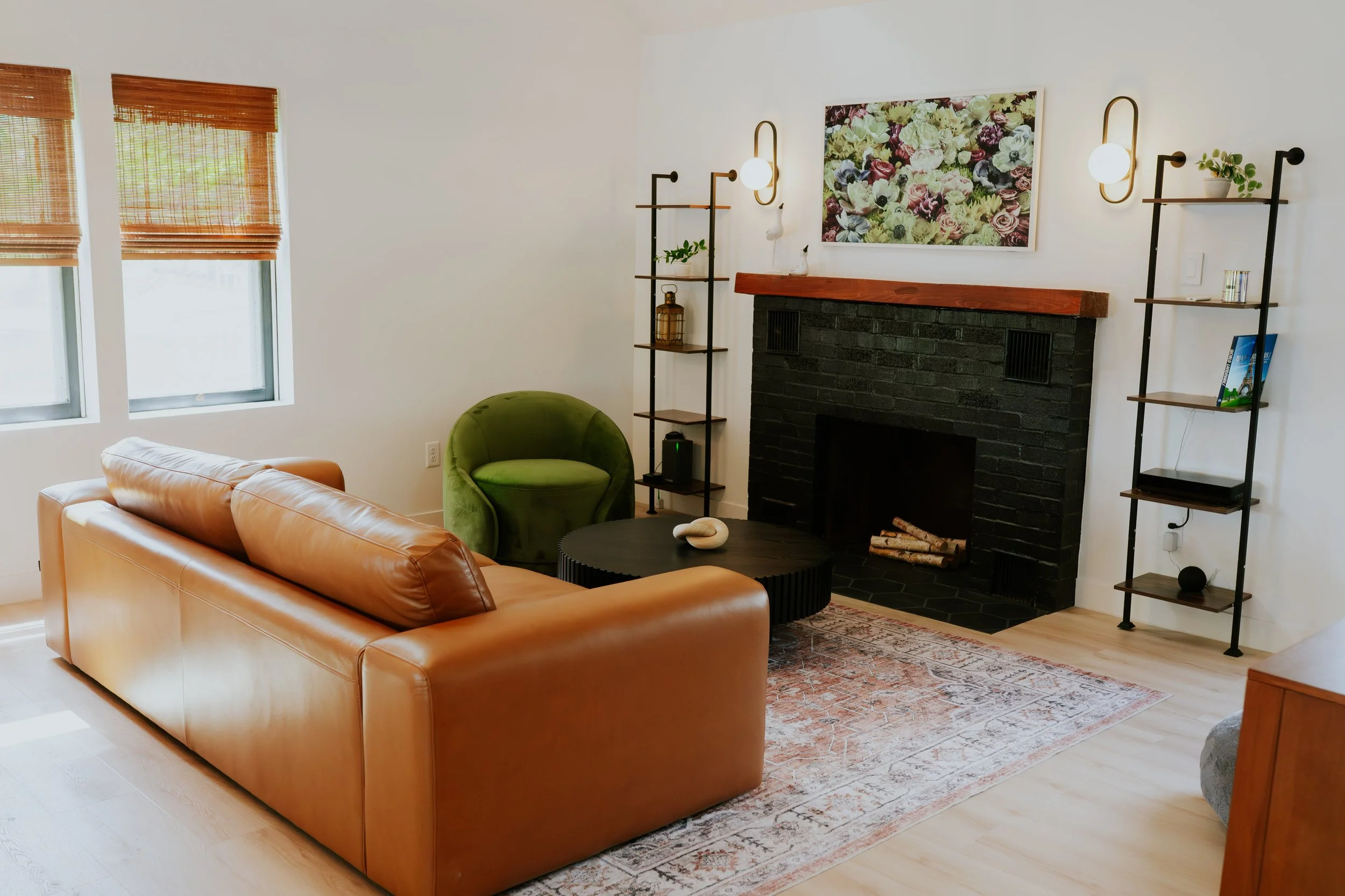 Living room with tan leather sofa, green velvet armchair, black round coffee table, fireplace with black brick and wooden mantel, floral artwork above fireplace, two wall sconces, black metal shelves with decorative items, bamboo window blinds, hardwood floor, area rug.