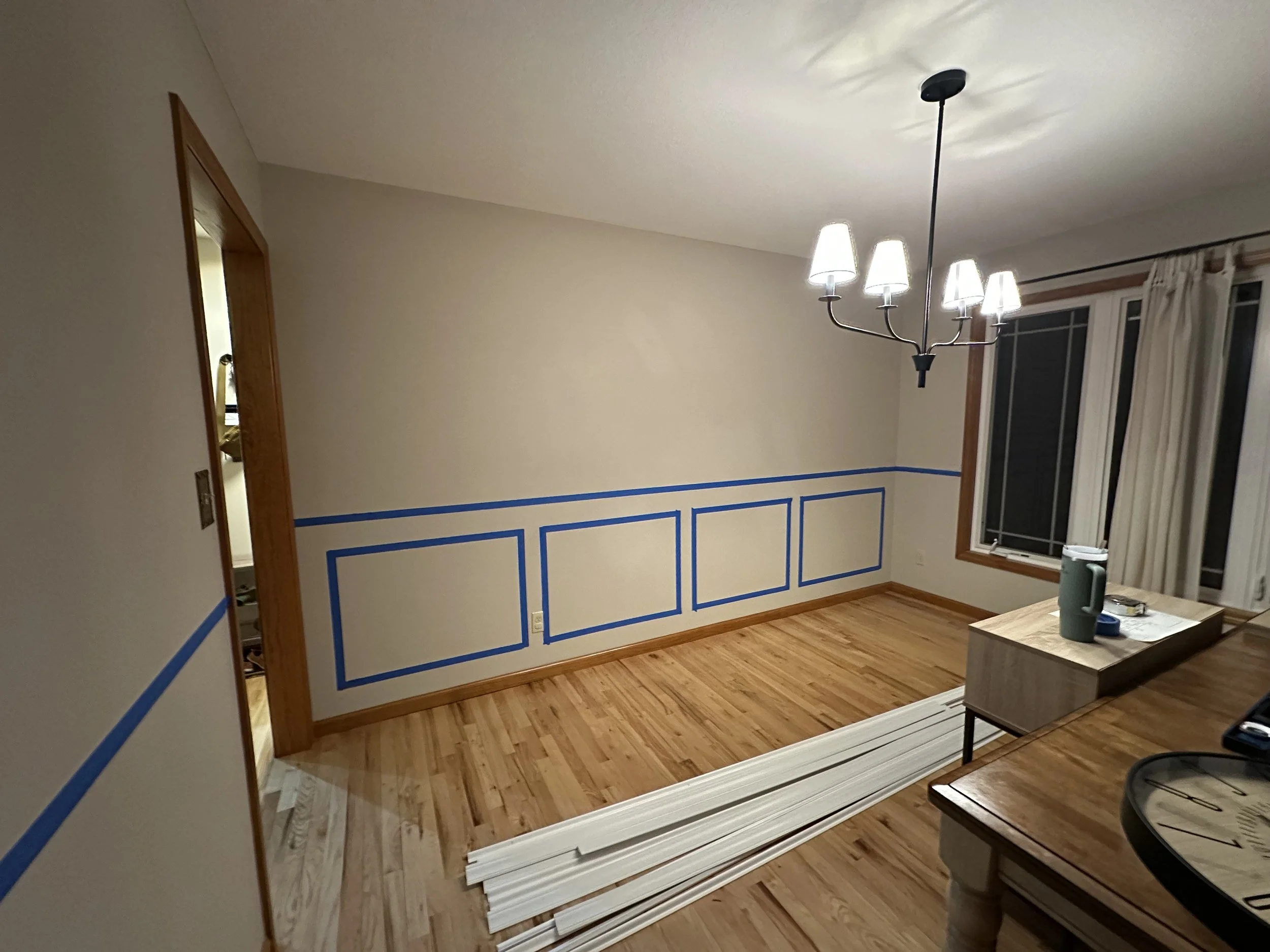 Empty dining room with hardwood floors, beige walls with blue paint trim, a window with white curtains, a black chandelier, and a wooden sideboard.