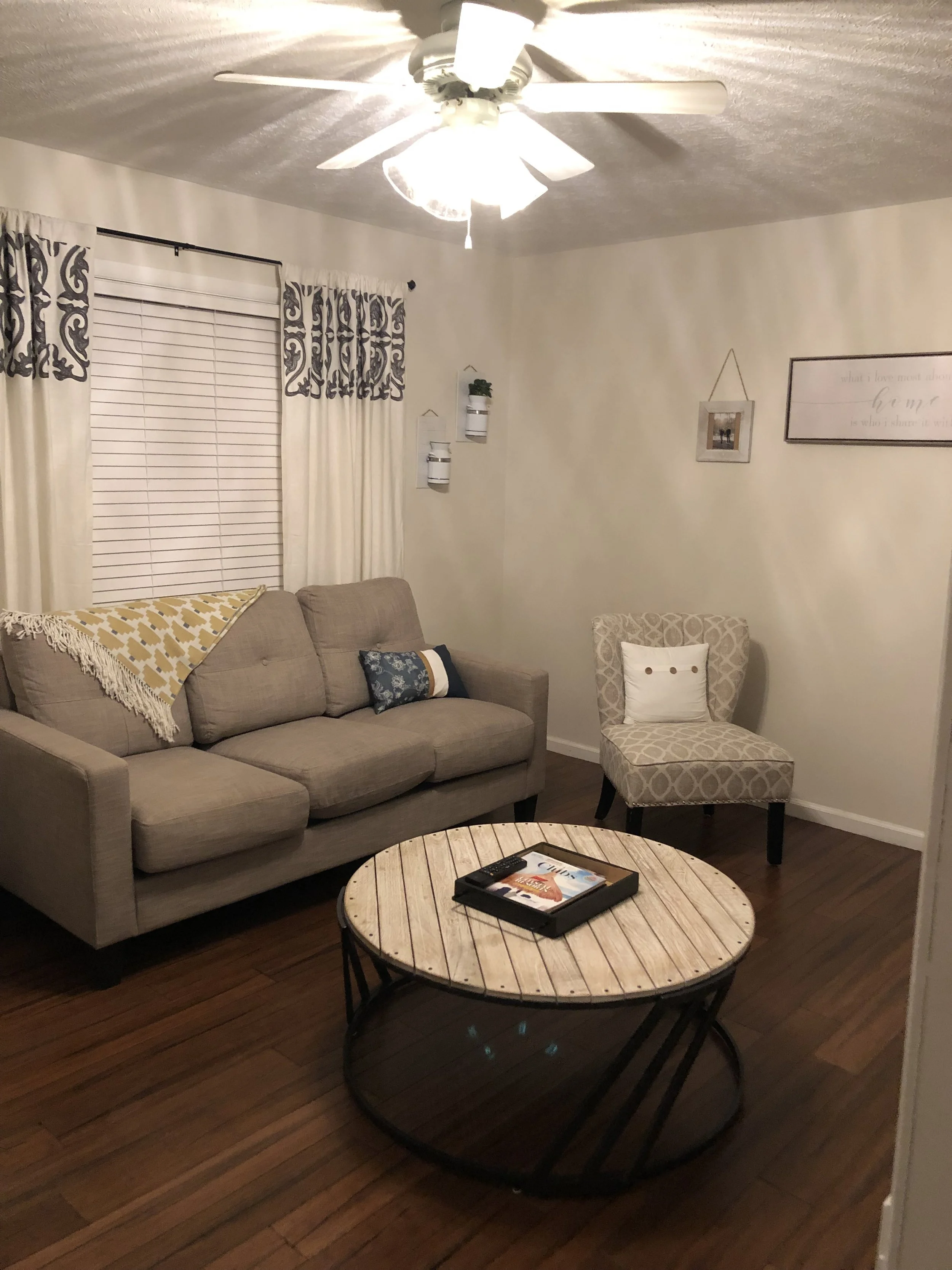 Living room with beige sofa, patterned armchair, round coffee table, ceiling fan, window with curtains, wall decorations, and hardwood floors.
