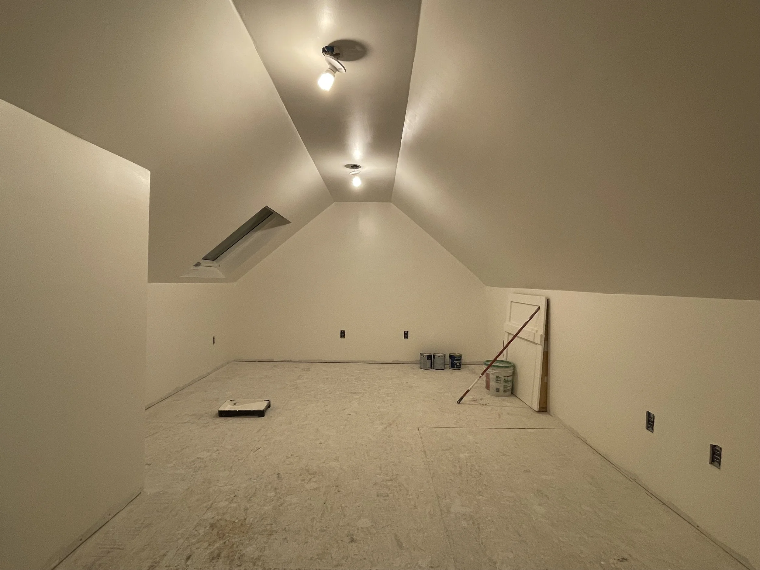 An unfinished attic room with sloped ceiling, painted white, and no flooring, showing drywall, with construction tools and supplies such as paint cans, a small step ladder, and a bucket, under ceiling lights.