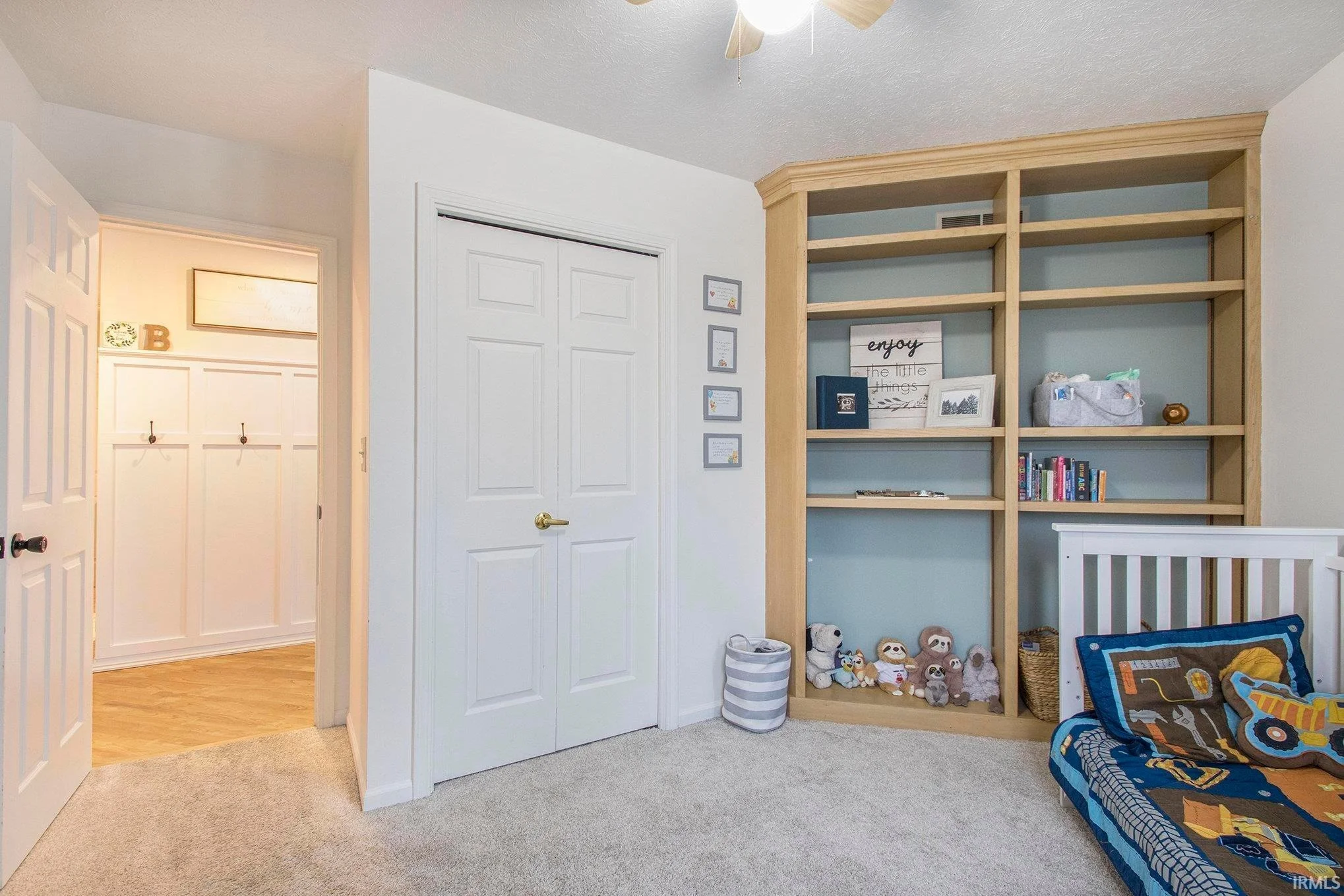 A children's bedroom featuring a partially open door, white walls, a built-in wooden bookshelf with various decorations and toys, a small bed with colorful bedding and pillows, and a laundry basket on the carpeted floor.
