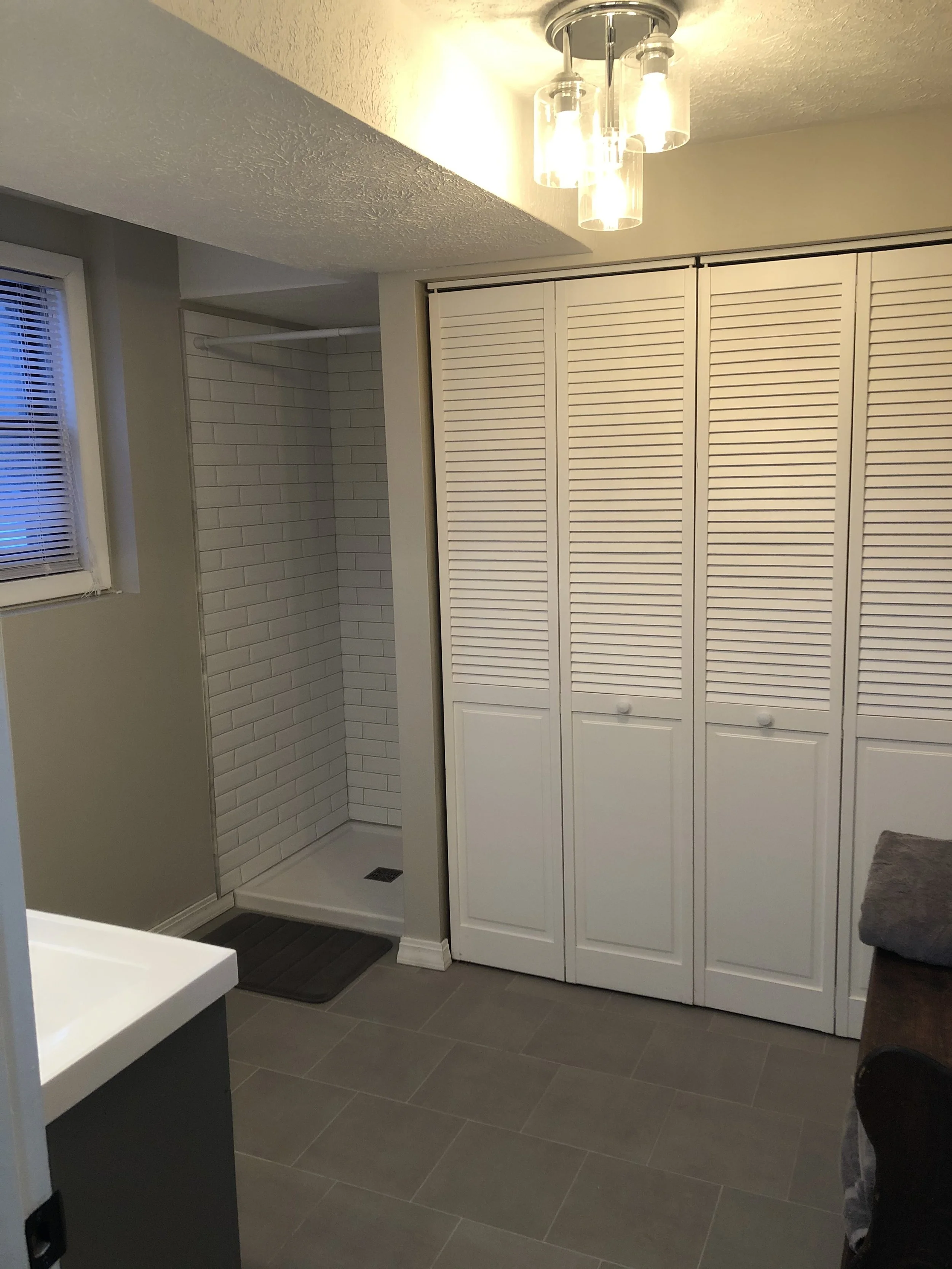 Bathroom with shower, window with blinds, louvered closet doors, gray tile floor, and ceiling light fixture.
