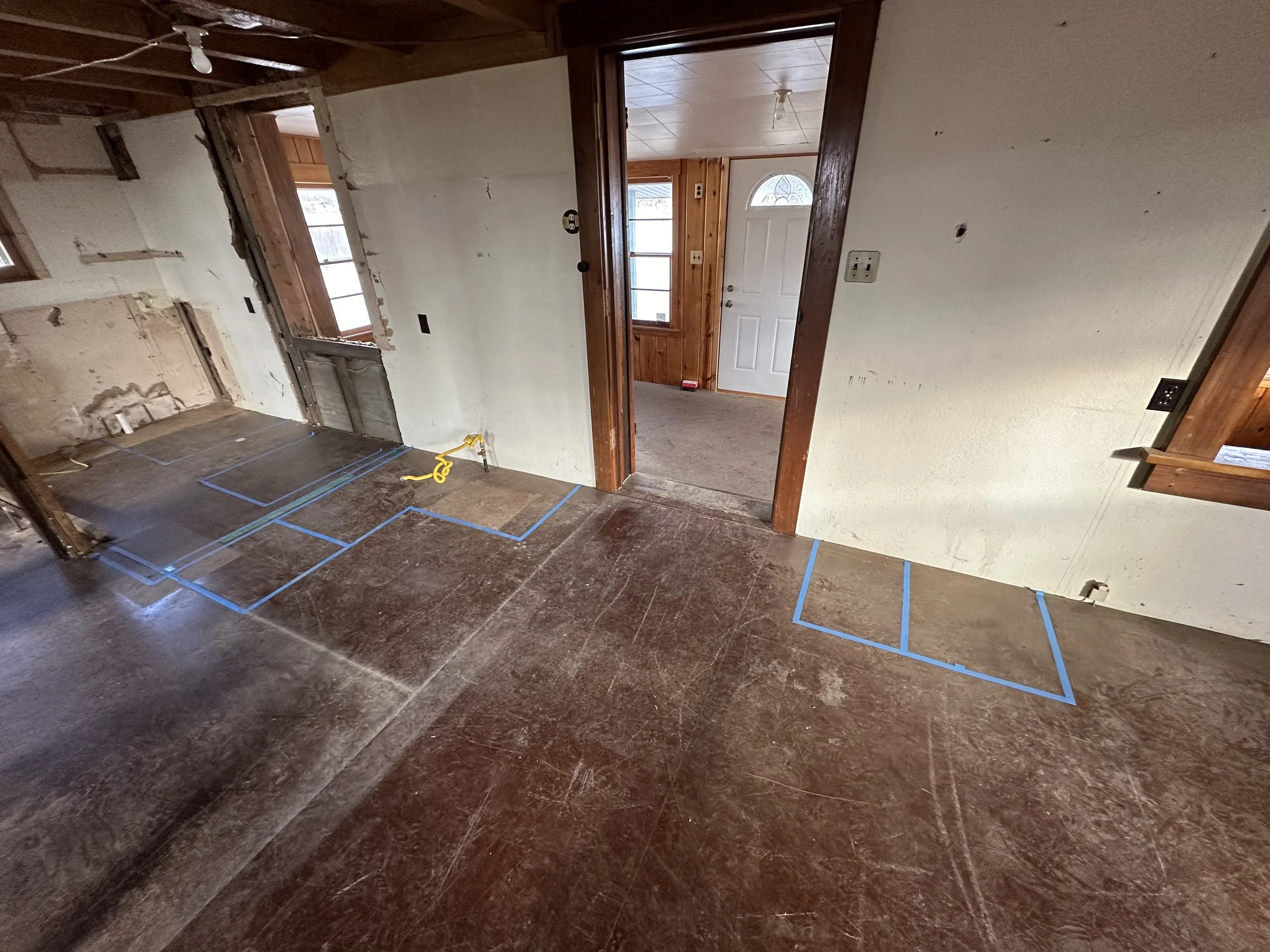 Interior room undergoing renovation with marked areas on the floor, some exposed walls and ceiling, and a doorway leading to a small enclosed porch area with a white door.